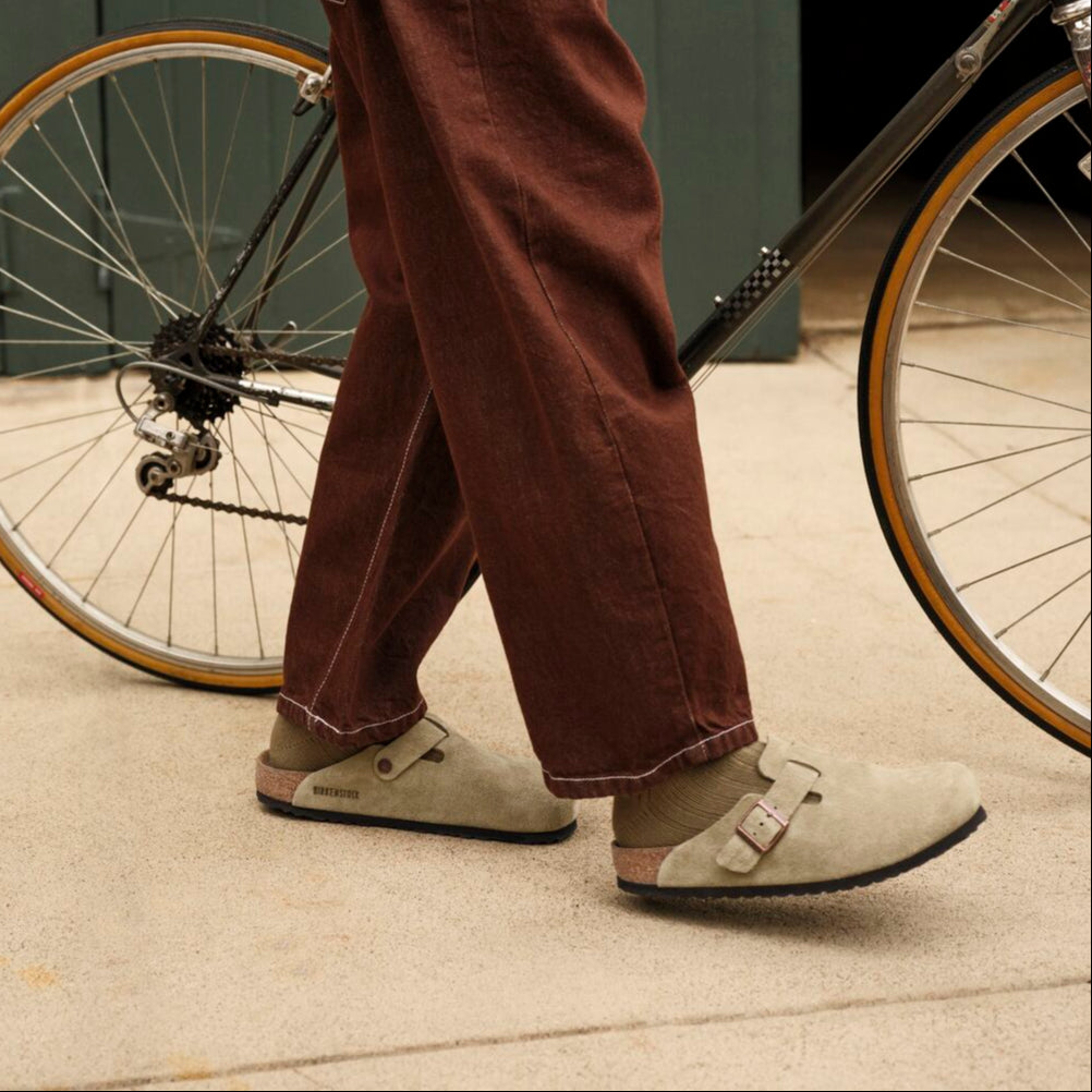 Person wearing brown pants and a green jacket standing next to a bicycle indoors.
