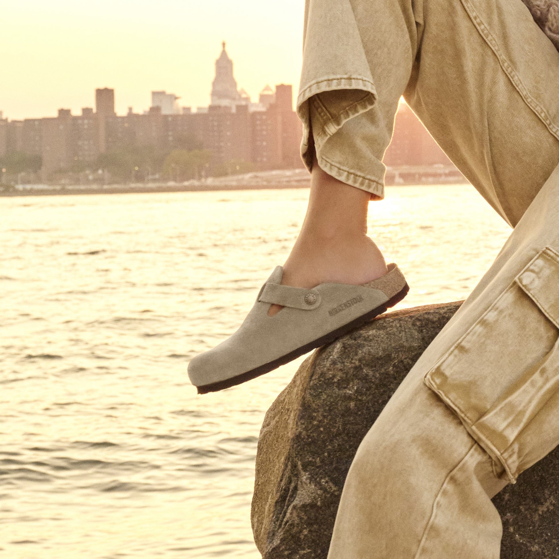 Person wearing beige clogs standing on a rock by a waterfront with a city skyline in the background.