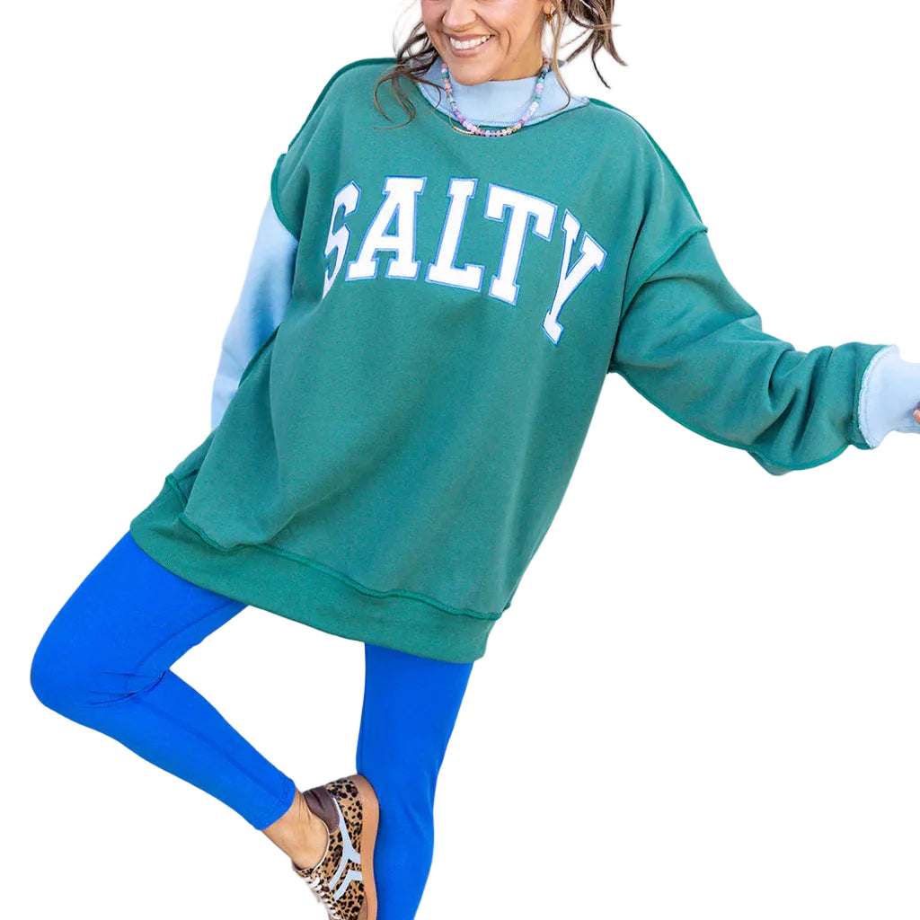 Woman wearing a green sweatshirt with 'SALTY' text, blue leggings, and leopard print shoes against a white background.