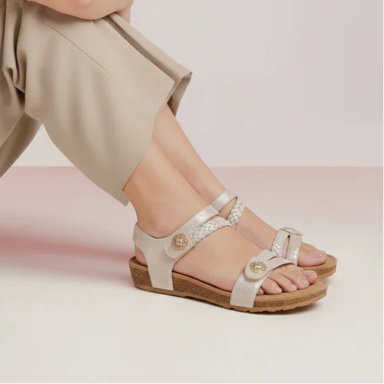 Woman sitting on a white surface wearing beige pants and sandals against a light pink background