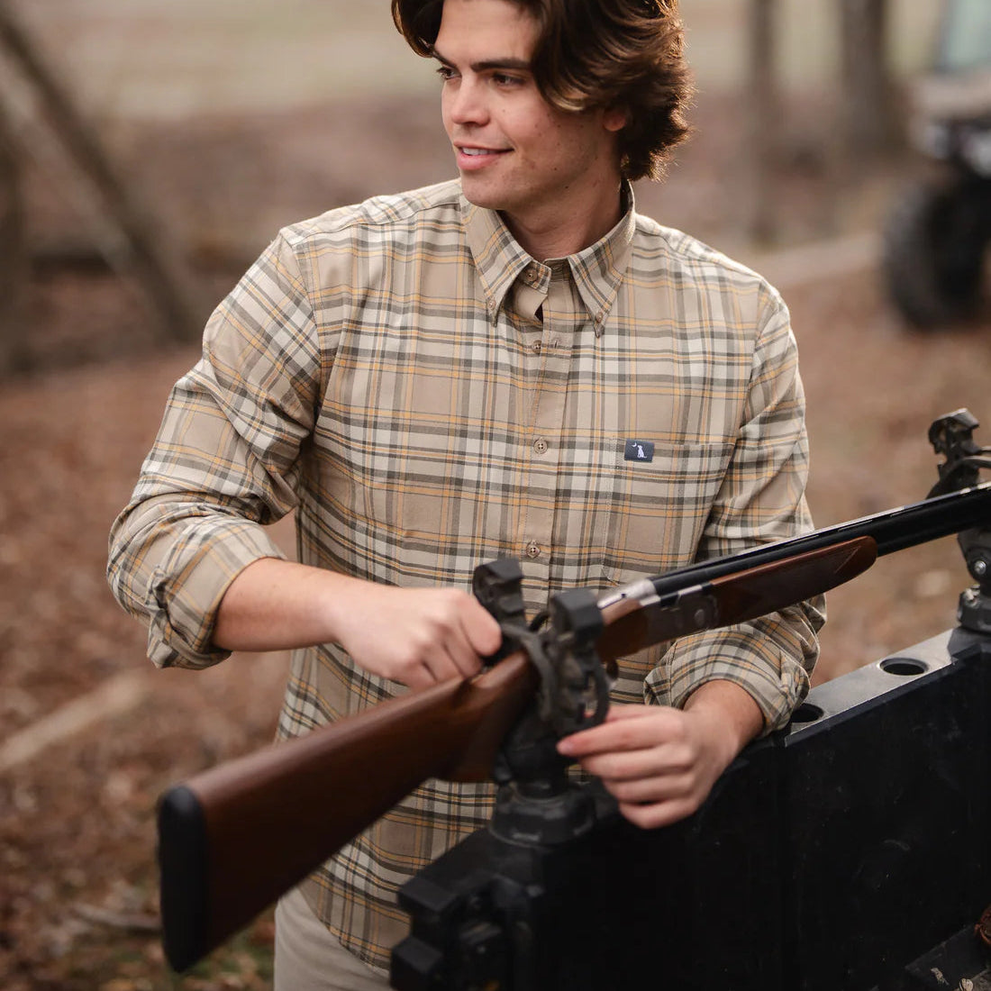 Man holding a rifle outdoors with trees and a field in the background