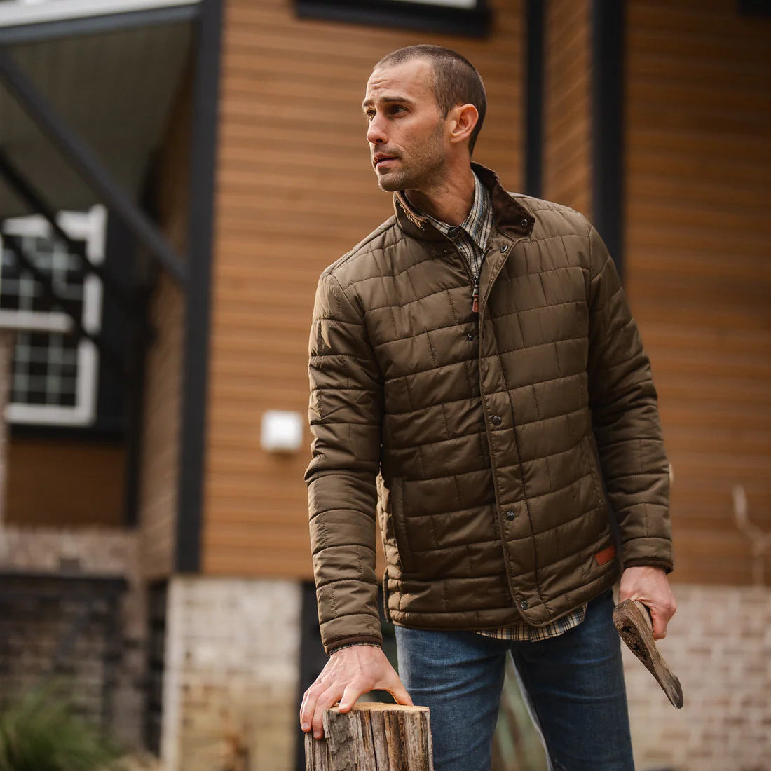 Man in a brown quilted jacket standing next to a wooden stump outdoors.