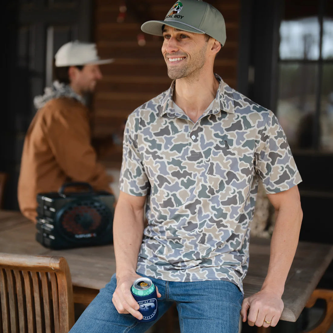 Man in a patterned shirt and cap holding a can, standing in a rustic setting with another person in the background.