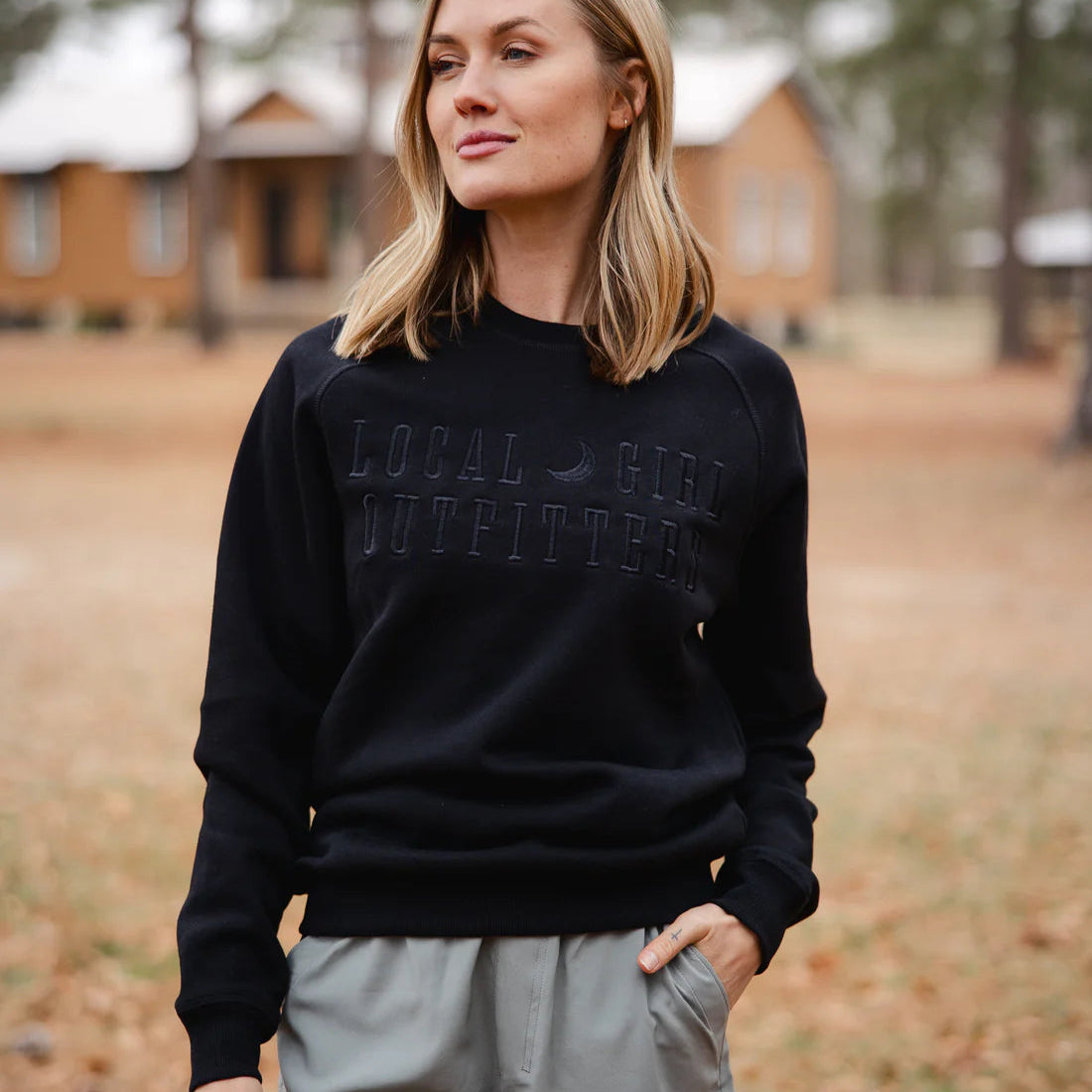 Woman wearing a black sweatshirt with text, standing in a field with trees and a building in the background.