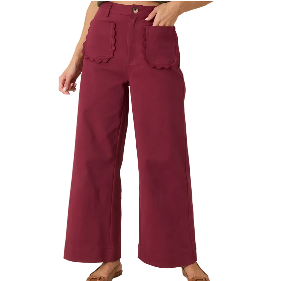 A person wearing high-waisted wide-leg pants in a deep burgundy or maroon color, featuring scalloped edges on the front patch pockets and a button closure, paired with tan flat shoes against a plain beige studio background.