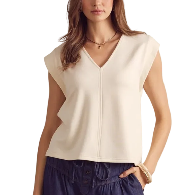 A woman modeling a sleeveless cream knit top with a V-neckline and front zipper, paired with dark blue pants, standing against a light beige brick wall.