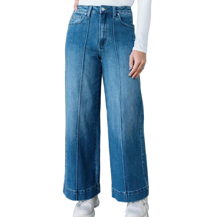 A person wearing blue wide-leg denim jeans with a straight-to-flared leg and cuffed hems, paired with a white long-sleeve top and white sneakers, standing against a white brick wall.