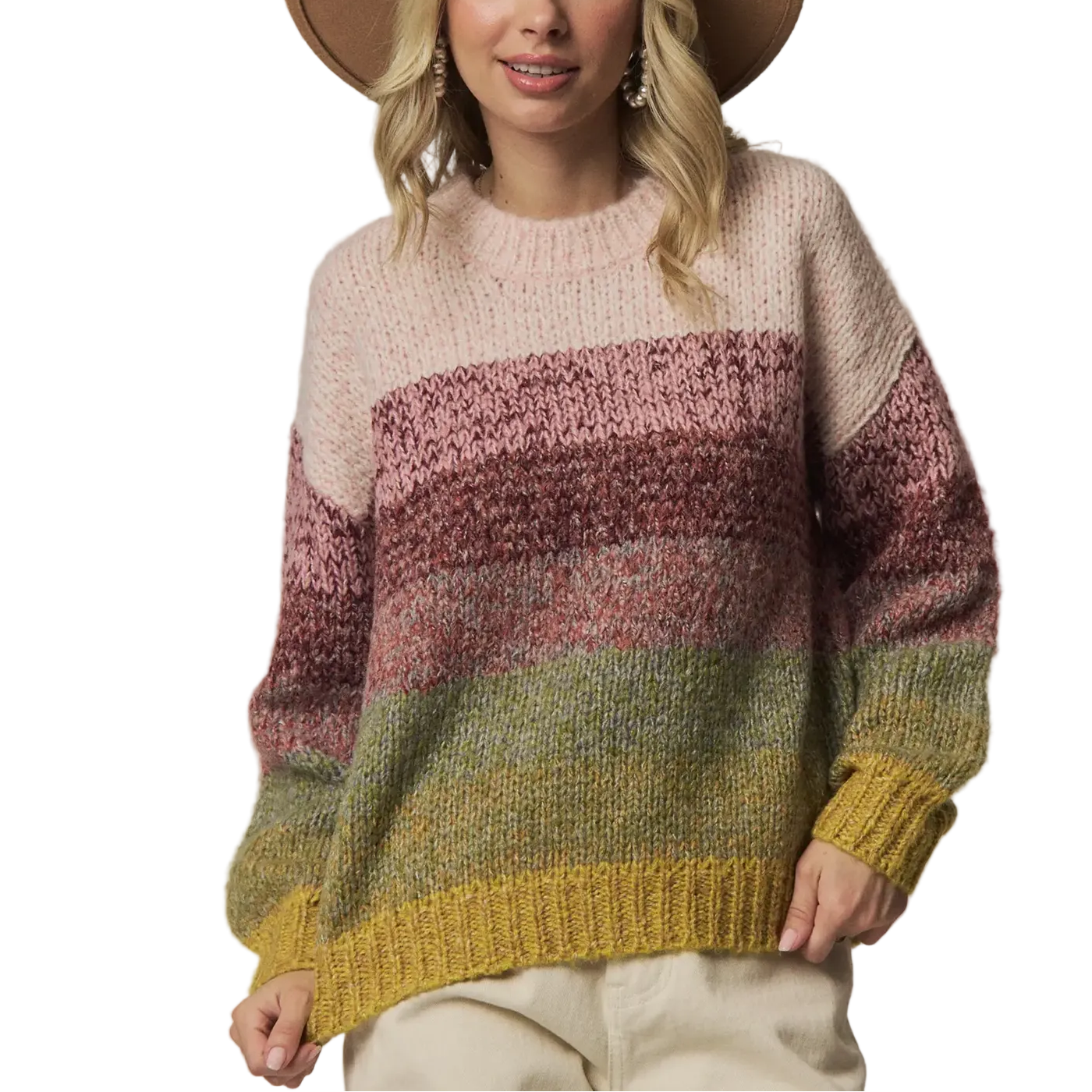 A woman wearing a chunky knit striped sweater with horizontal bands of cream, dusty rose, mauve, sage green, and mustard yellow, paired with a wide-brimmed tan hat and light beige pants, against a solid light gray studio background.