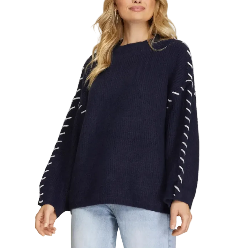 A woman wearing a navy blue ribbed knit sweater with white dashed line patterns on the sleeves, paired with light blue jeans, against a solid gray studio background.